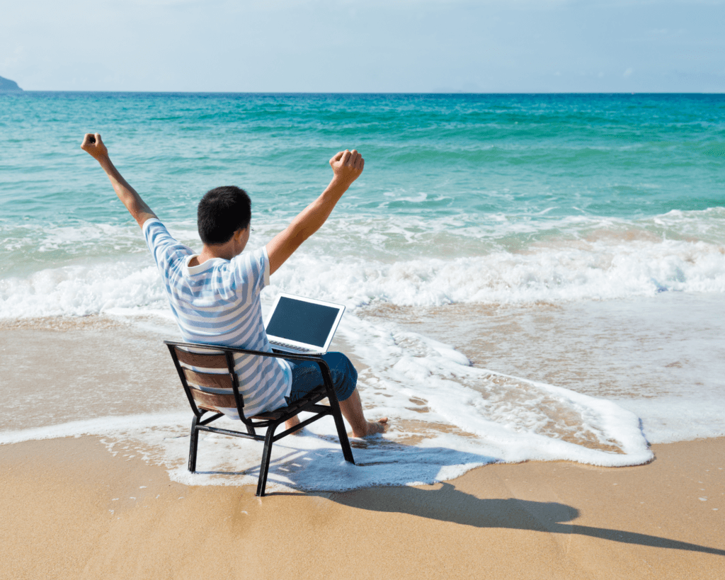 Un homme assis sur une chaise en bois, en bord de mer, il lève les bras de joie avec son PC sur les genoux 